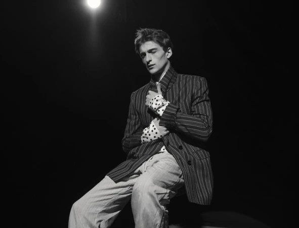 Male model seated in a dark pinstripe jacket and polka dot shirt under dramatic spotlight against a black background.