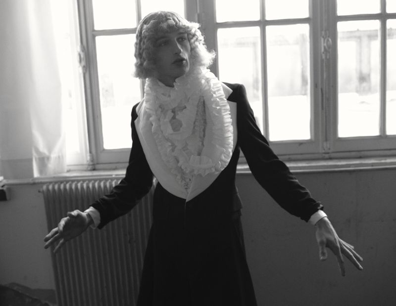 Vasko Luyckx wearing a black tailored jacket, white ruffled collar, and styled wig in front of large multi-pane windows.