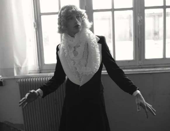 Vasko Luyckx wearing a black tailored jacket, white ruffled collar, and styled wig in front of large multi-pane windows.