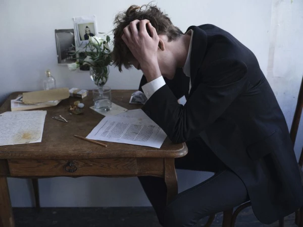 A man in a black suit sits slumped over a wooden desk covered with papers and flowers, resting his head in his hand.