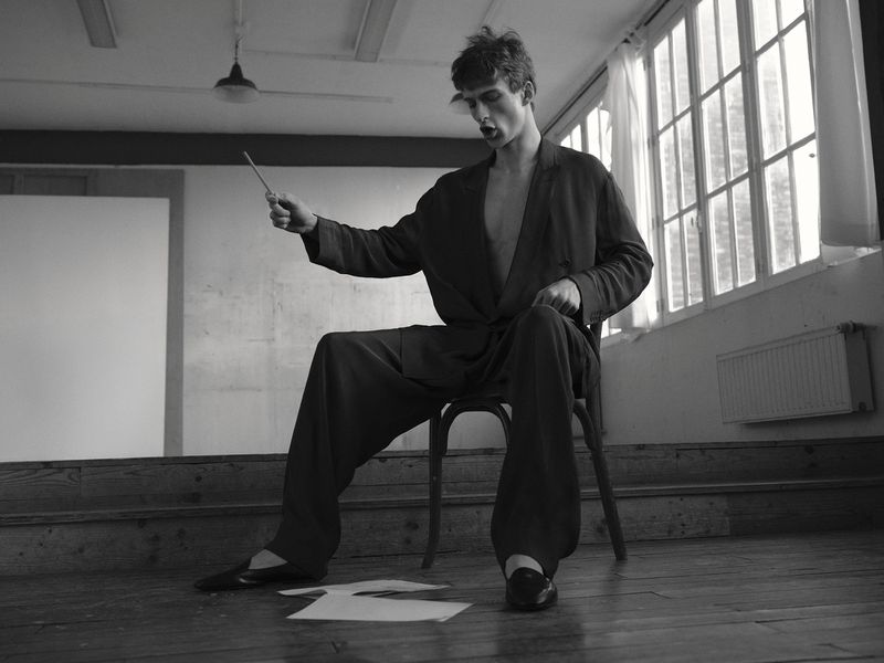 Model Vasko Luyckx seated on a wooden chair in an oversized dark suit holding a baton in a monochrome room with large windows.