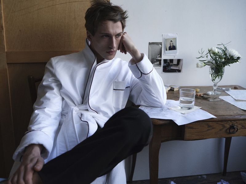 Model Vasko Luyckx wearing a white robe with black piping seated at a wooden desk with flowers and handwritten papers.