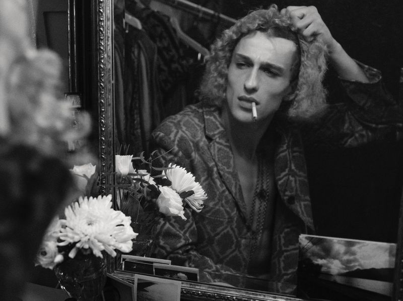 Black and white mirror reflection of a curly-haired man smoking a cigarette in a patterned jacket beside white flowers.