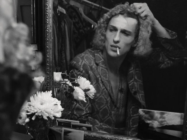 Black and white mirror reflection of a curly-haired man smoking a cigarette in a patterned jacket beside white flowers.