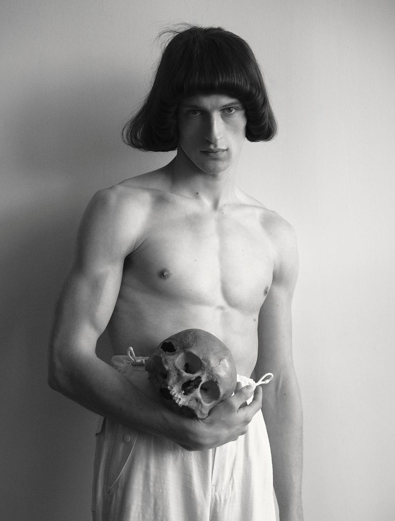 Shirtless young man with a blunt bob haircut holding a human skull at his waist against a plain studio backdrop.