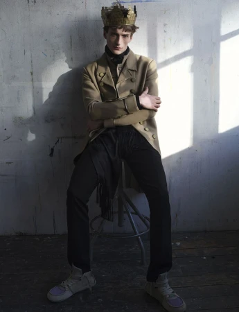 Model Vasko Luyckx wearing a gold crown and double-breasted tan coat seated on a stool against a sunlit wall.