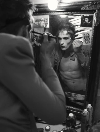 Black and white portrait of a man applying makeup with a brush while looking into a dressing room mirror.