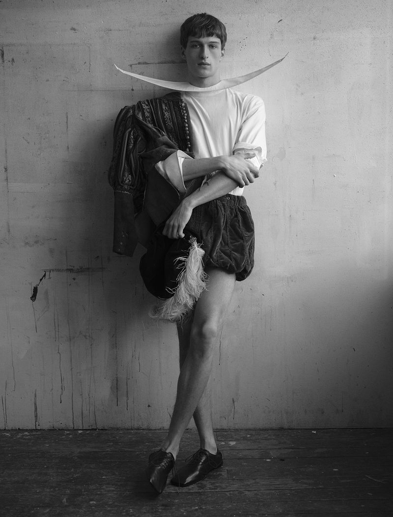 Black and white full-length portrait of a man in theatrical Elizabethan attire, a wide crescent collar, and puffed shorts against a wall.