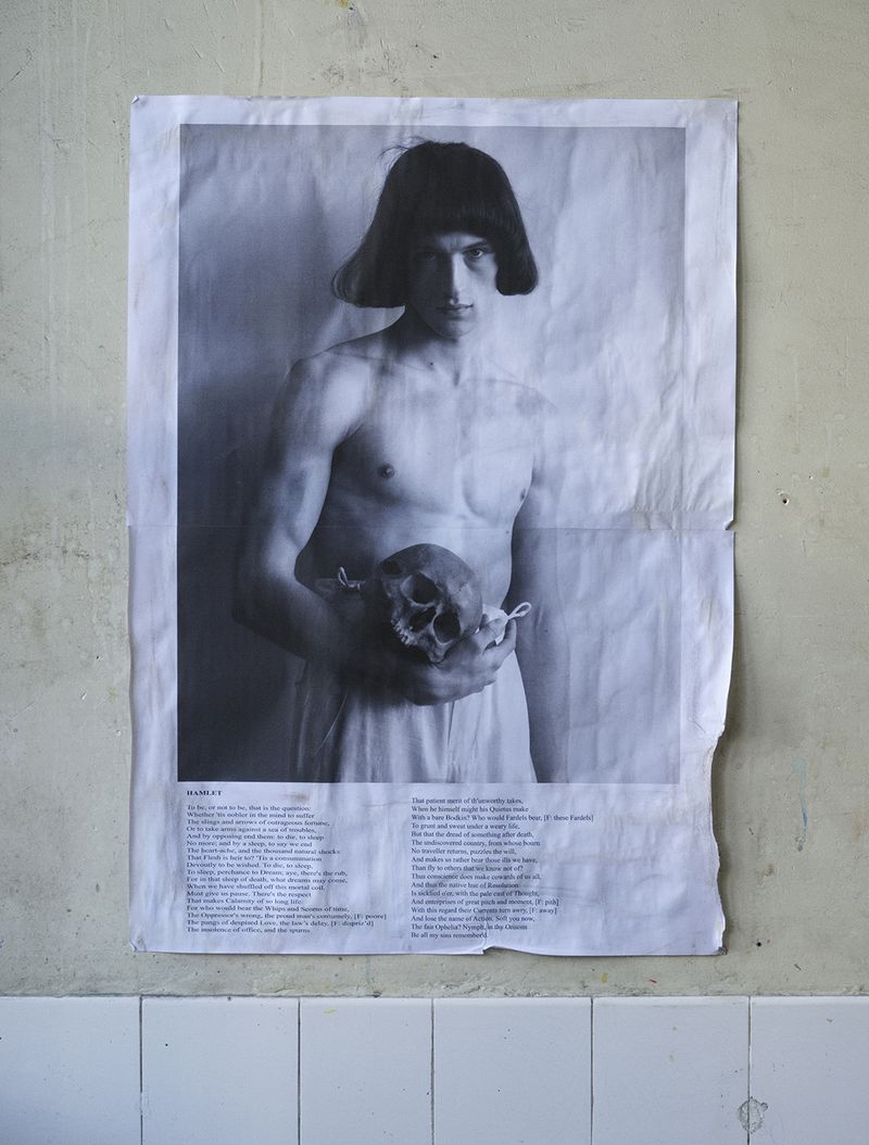 Monochrome poster of a shirtless man with dark hair holding a skull, taped to a distressed wall above white tiles.