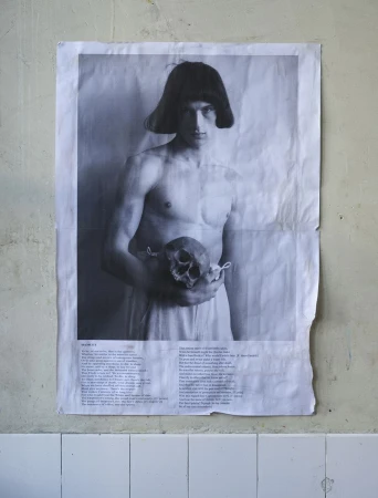Monochrome poster of a shirtless man with dark hair holding a skull, taped to a distressed wall above white tiles.