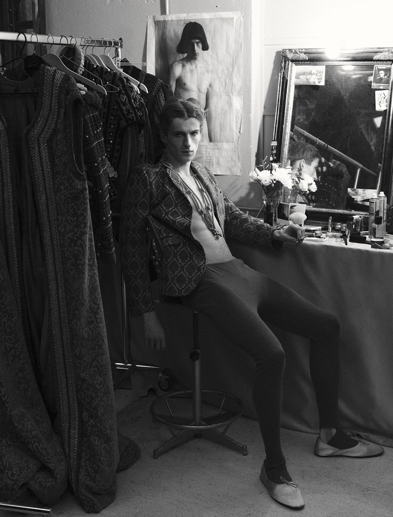 Model Vasko Luyckx seated backstage in a dressing room wearing a patterned jacket and tights for Dapper Dan Magazine.
