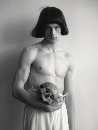 Shirtless young man with a blunt bob haircut holding a human skull at his waist against a plain studio backdrop.