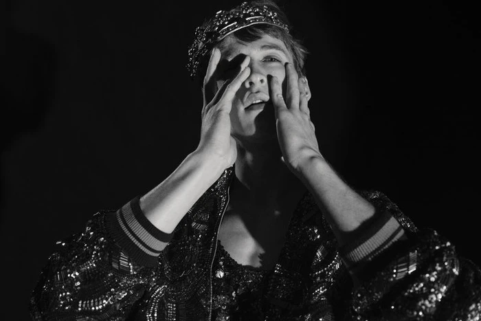 Model Vasko Luyckx wearing a beaded crown and sequined jacket with hands to his face against a black backdrop.