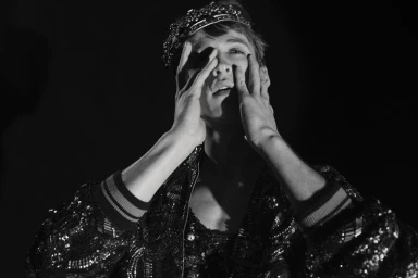Model Vasko Luyckx wearing a beaded crown and sequined jacket with hands to his face against a black backdrop.