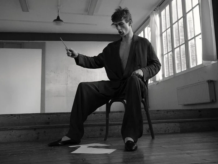 Model Vasko Luyckx seated on a wooden chair in an oversized dark suit holding a baton in a monochrome room with large windows.