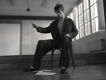 Model Vasko Luyckx seated on a wooden chair in an oversized dark suit holding a baton in a monochrome room with large windows.