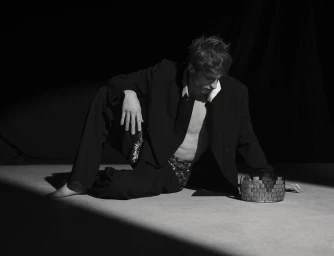 Model Vasko Luyckx sitting barefoot on a dark studio floor in an unbuttoned black suit jacket beside a metallic crown.