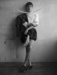 Black and white full-length portrait of a man in theatrical Elizabethan attire, a wide crescent collar, and puffed shorts against a wall.