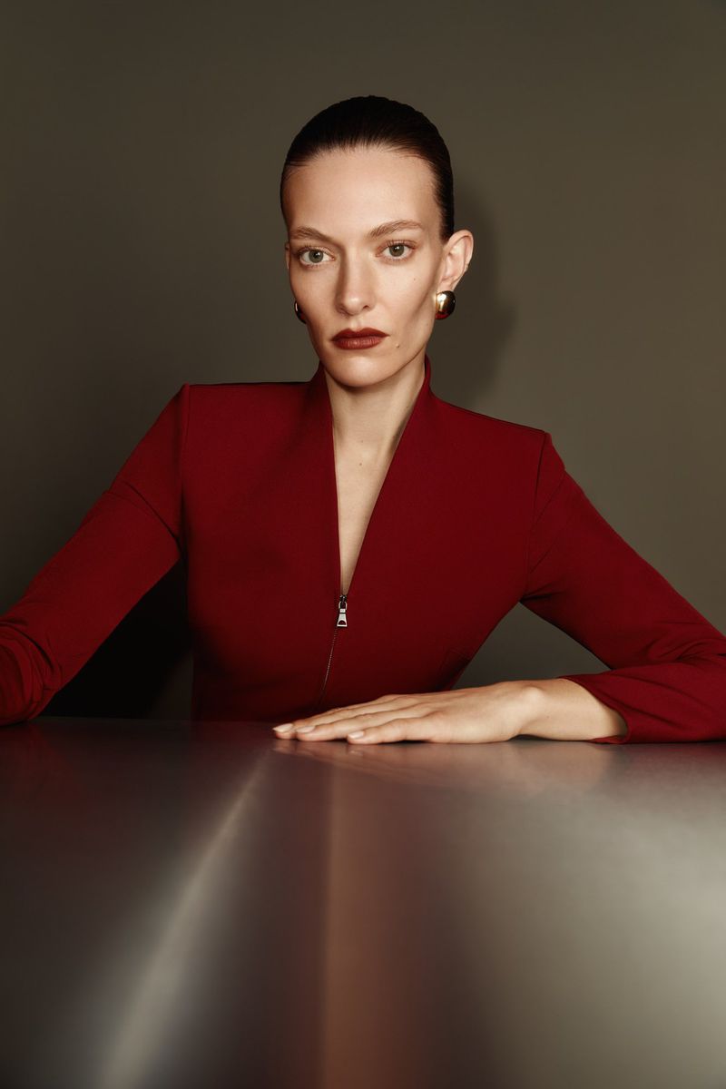Portrait of a woman with slicked-back hair, wearing a red suit with zipper, gold earrings, and dark red lipstick, sitting at a table.