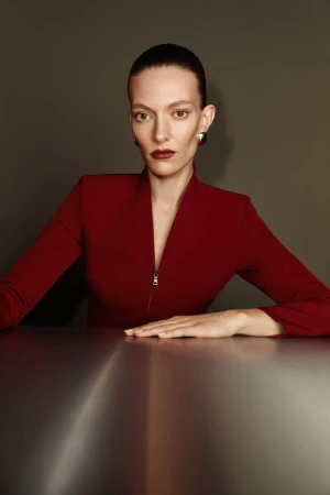 Portrait of a woman with slicked-back hair, wearing a red suit with zipper, gold earrings, and dark red lipstick, sitting at a table.