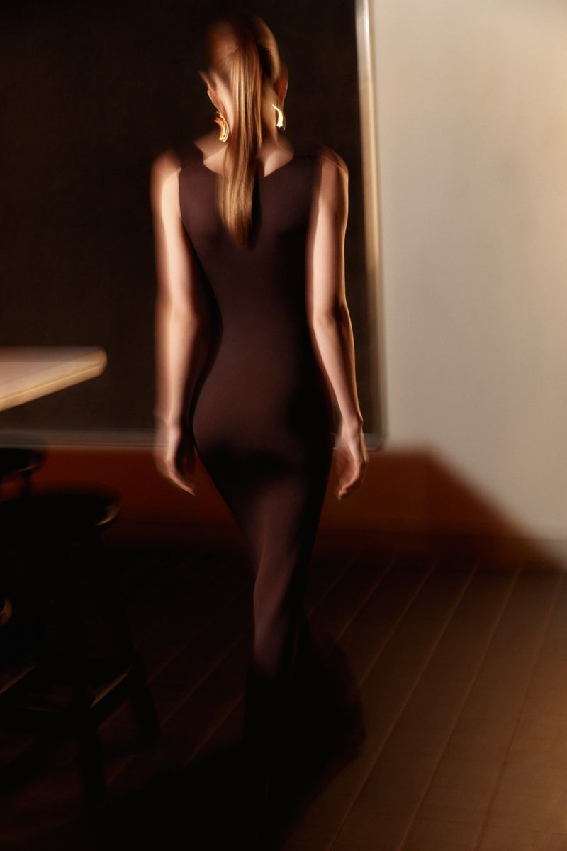 Elegant woman in a brown dress walking in a dimly lit room, showcasing a blend of fashion and interior design.