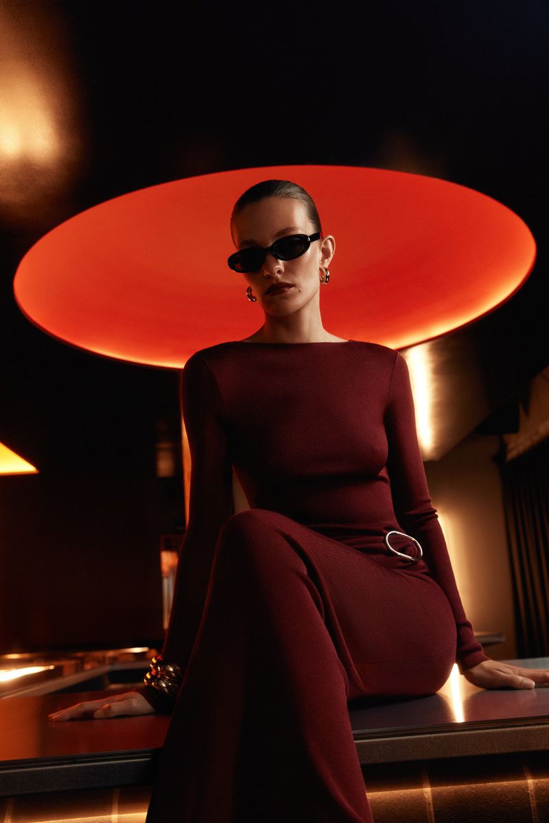 Stylish woman in burgundy dress and sunglasses sitting on a bar counter with vibrant orange lighting in the background, fashion shot.