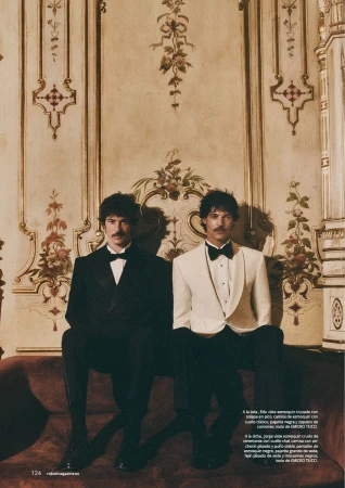 Edu Román and Jorge Roman seated in black and white tuxedos against an ornate classical wall with gilded mouldings.