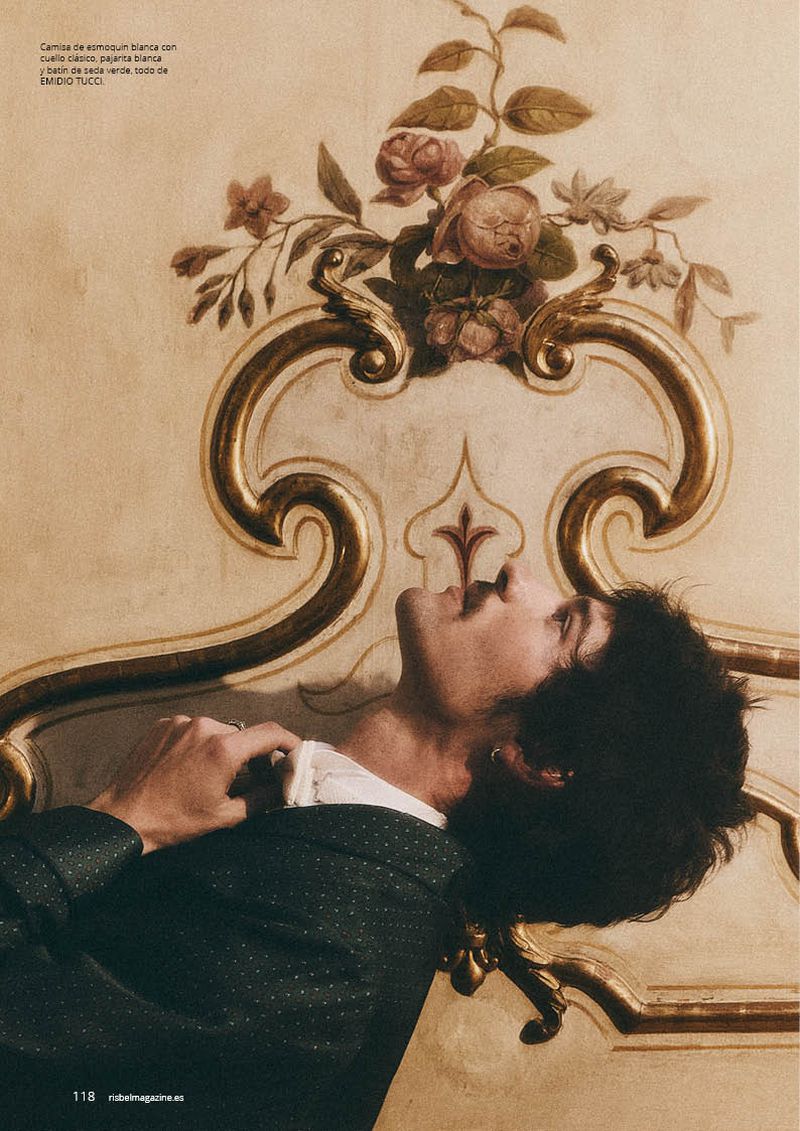Man in a dark tuxedo jacket leaning back against an ornate gilded wall with painted floral details in profile view.