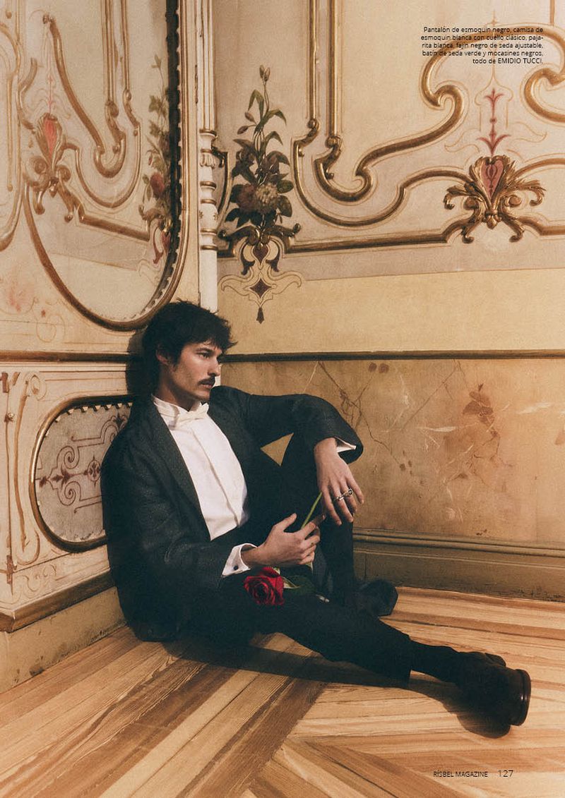 Man in a dark suit and white shirt sitting on a parquet floor holding a red rose against an ornate wall