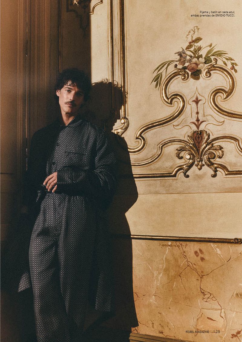 A male model with a moustache leans against an ornate gilded wall wearing a dark patterned silk pajama suit and robe.