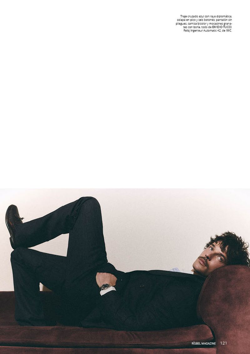 A man in a dark pinstripe suit reclines across a deep burgundy velvet sofa against a plain white wall in an editorial layout.