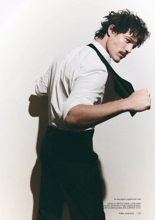 Model Jorge Román wearing a white dress shirt and black trousers with an untied black tie against a white background.