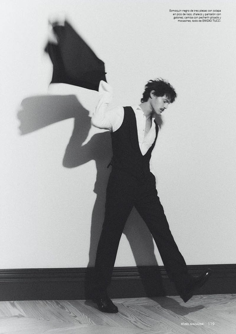 Man wearing a black vest and trousers taking off a jacket against a plain wall in an editorial photograph