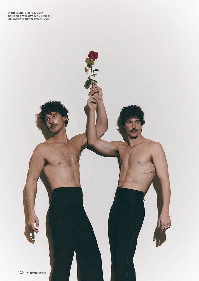 Two shirtless men with mustaches wearing high-waisted black trousers holding a single red rose together against a light background.