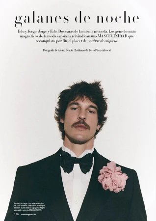 Man with a mustache wearing a black tuxedo jacket with a pink flower lapel pin and black bow tie against a white background.