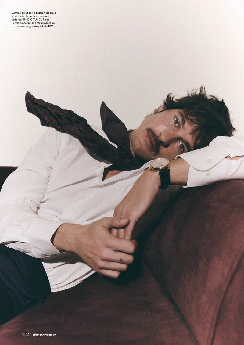 A man with a mustache lying back on a burgundy sofa wearing a white dress shirt and a dark silk neckerchief.