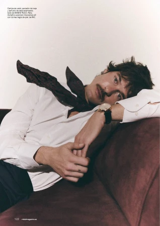 A man with a mustache lying back on a burgundy sofa wearing a white dress shirt and a dark silk neckerchief.