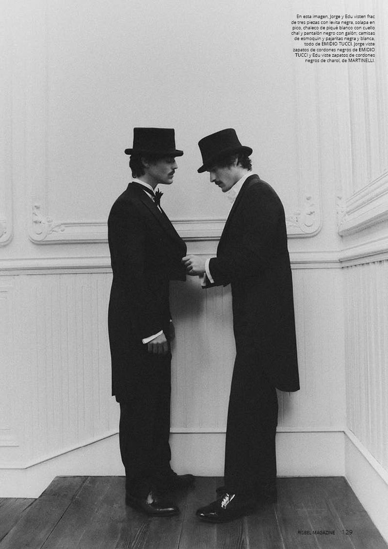 Two men wearing black formal tailcoats and top hats facing each other against a white wall with wainscoting.
