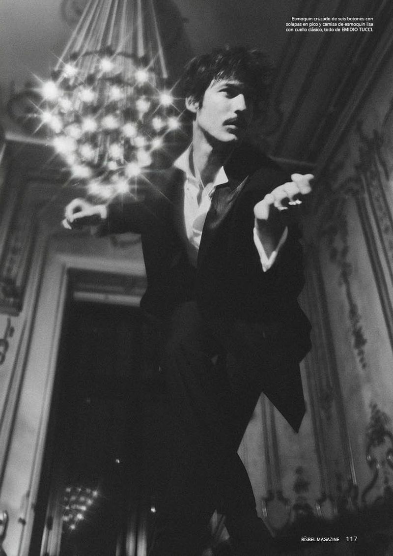 Man in a black tuxedo and open white shirt poses dynamically beneath a glowing crystal chandelier in an ornate hall.