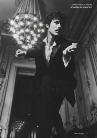 Man in a black tuxedo and open white shirt poses dynamically beneath a glowing crystal chandelier in an ornate hall.