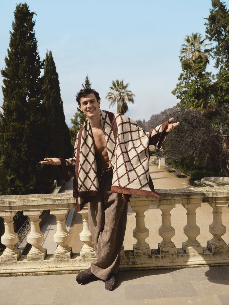 Model Xavier Serrano smiling with open arms wearing a patterned poncho and brown trousers on a stone balustrade terrace.