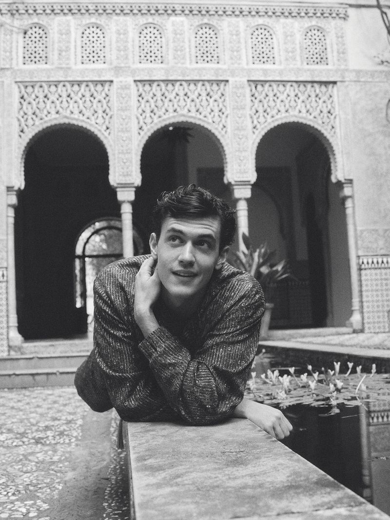 Model Xavier Serrano leaning over a stone ledge in front of ornate Moorish arches in a black and white fashion portrait.