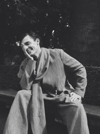 Xavier Serrano wearing a textured draped suit and matching scarf while seated smiling on a stone bench in a park.