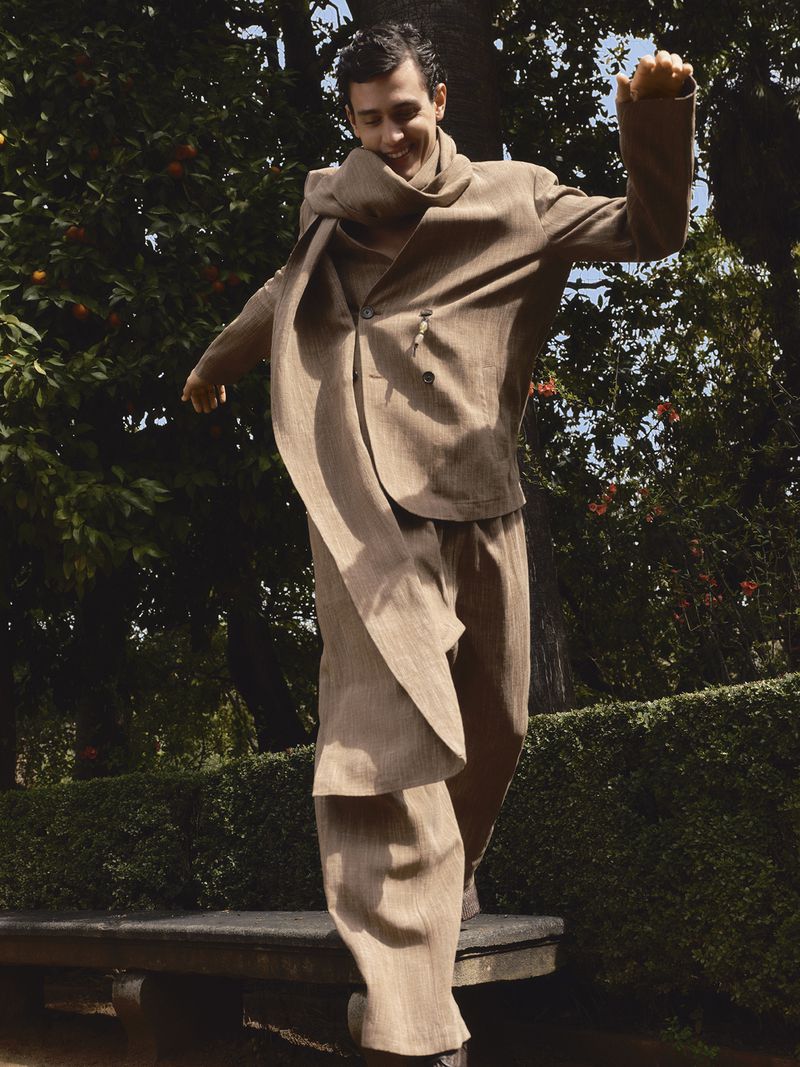 Man in tan suit balances on bench in park-like setting, adorned with trees, enjoying natural light and an upbeat mood, showcasing fashion.