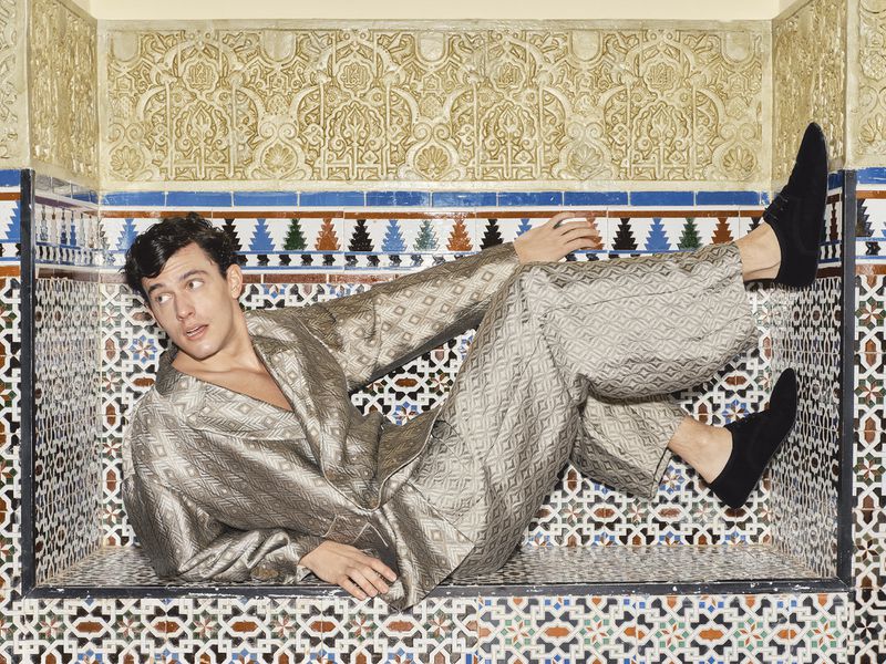 Model Xavier Serrano reclining inside an ornate geometric tiled alcove wearing a patterned metallic jacquard suit and black shoes.