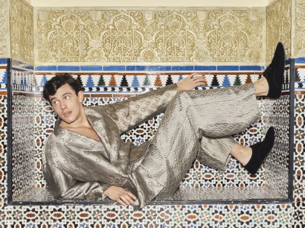 Model Xavier Serrano reclining inside an ornate geometric tiled alcove wearing a patterned metallic jacquard suit and black shoes.