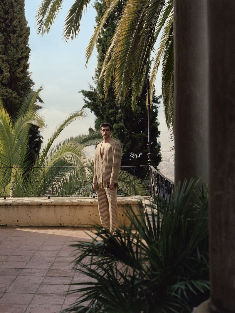 Model Xavier Serrano wearing a beige textured suit standing on a sunlit tiled terrace overlooking palm trees and landscape.