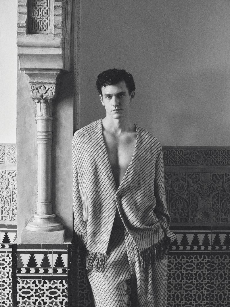 Monochrome portrait of model Xavier Serrano in a striped fringe jacket leaning against an ornate carved architectural pillar.