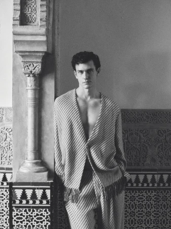 Monochrome portrait of model Xavier Serrano in a striped fringe jacket leaning against an ornate carved architectural pillar.