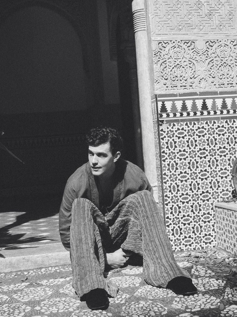 Model Xavier Serrano seated on the ground beside ornate mosaic tilework in a black and white editorial portrait.